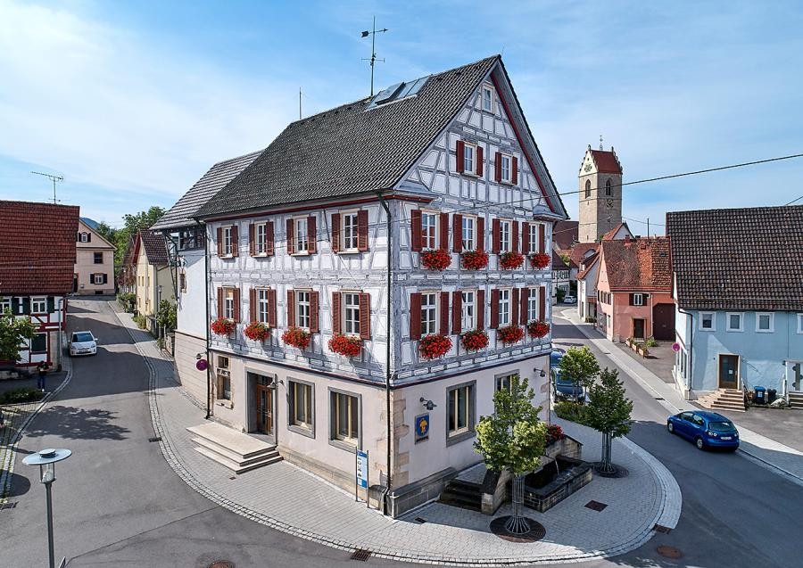 Dreistöckiges Gebäude mit Sockelgeschoss und zwei Fachwerkgeschossen, Satteldach, Blumenschmuck, Schild mit dem Wappen von Ofterdingen., an der Straße weitere Häuser und ein Kirchturm.