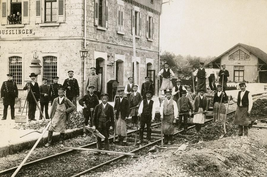 Teil des Bahnhofsgebäudes und etwas entfernt ein Schuppen, davor teilweise unvollständige Schienen, eine Gruppe von etwa zwanzig Männern in Dienstuniformen und Arbeitskleidung stehen vor dem Gebäude und im Gleisbett, aus dem Fenster schauen zwei Personen, historische Fotografie