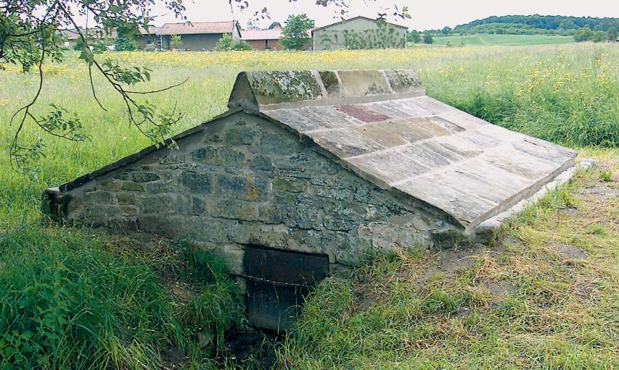 Niedriges über einem Graben errichtetes Steingebäude, in Satteldachform mit Steinplatten gedeckt, vorne ist eine Öffnung am Graben sichtbar